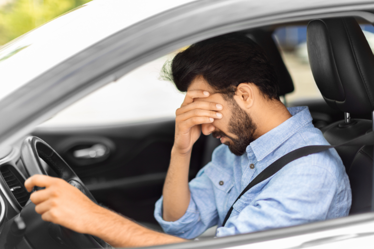 Homem dentro do carro com a mão no rosto, aparentando cansaço ou estresse no trânsito.
