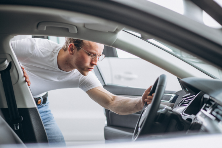 Homem inclinado para dentro de um carro, observando o interior do veículo e segurando o volante com uma das mãos.