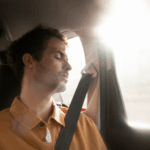 Homem sentado no banco traseiro de um carro, usando cinto de segurança, com a cabeça apoiada ao lado e luz natural entrando pela janela.