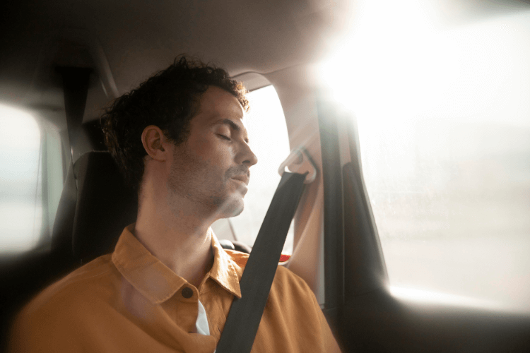Homem sentado no banco traseiro de um carro, usando cinto de segurança, com a cabeça apoiada ao lado e luz natural entrando pela janela.