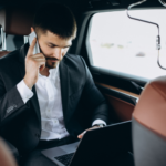 Executivo utilizando notebook e falando ao telefone no banco traseiro de um carro durante deslocamento corporativo.