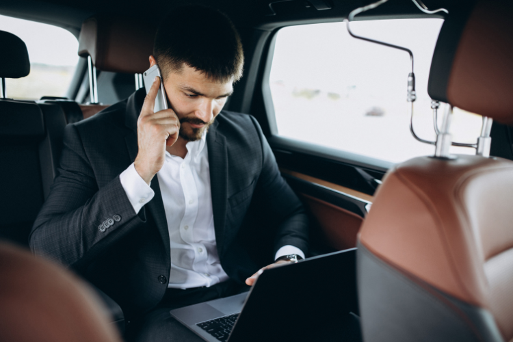 Executivo utilizando notebook e falando ao telefone no banco traseiro de um carro durante deslocamento corporativo.